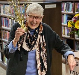 Carole Berard retirement photo. Carole is holding gold sparkle decoration and smiling in the library, with her left arm leaning on a display table with a sign that reads 'congratulations carole'.