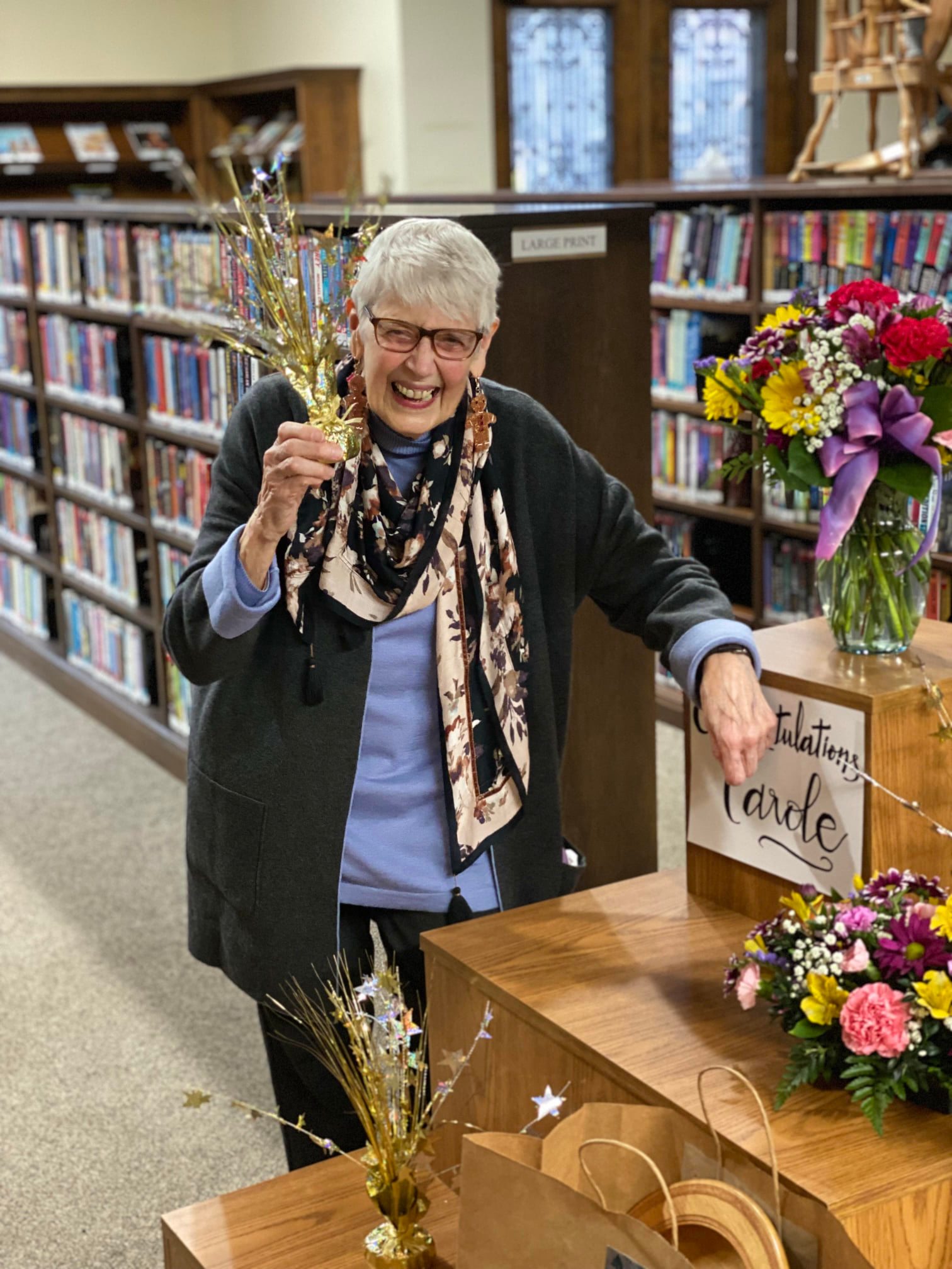 Carole Berard retirement photo. Carole is holding gold sparkle decoration and smilinf in the libary, with her left arm leaning on a display table w a sign that reads 'congratulations carole'.
