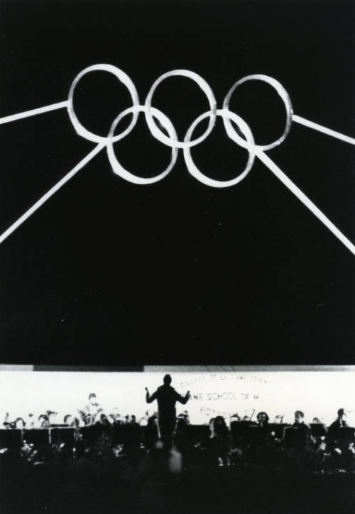 A conductor leads an orchestra beneath suspended Olympic rings.