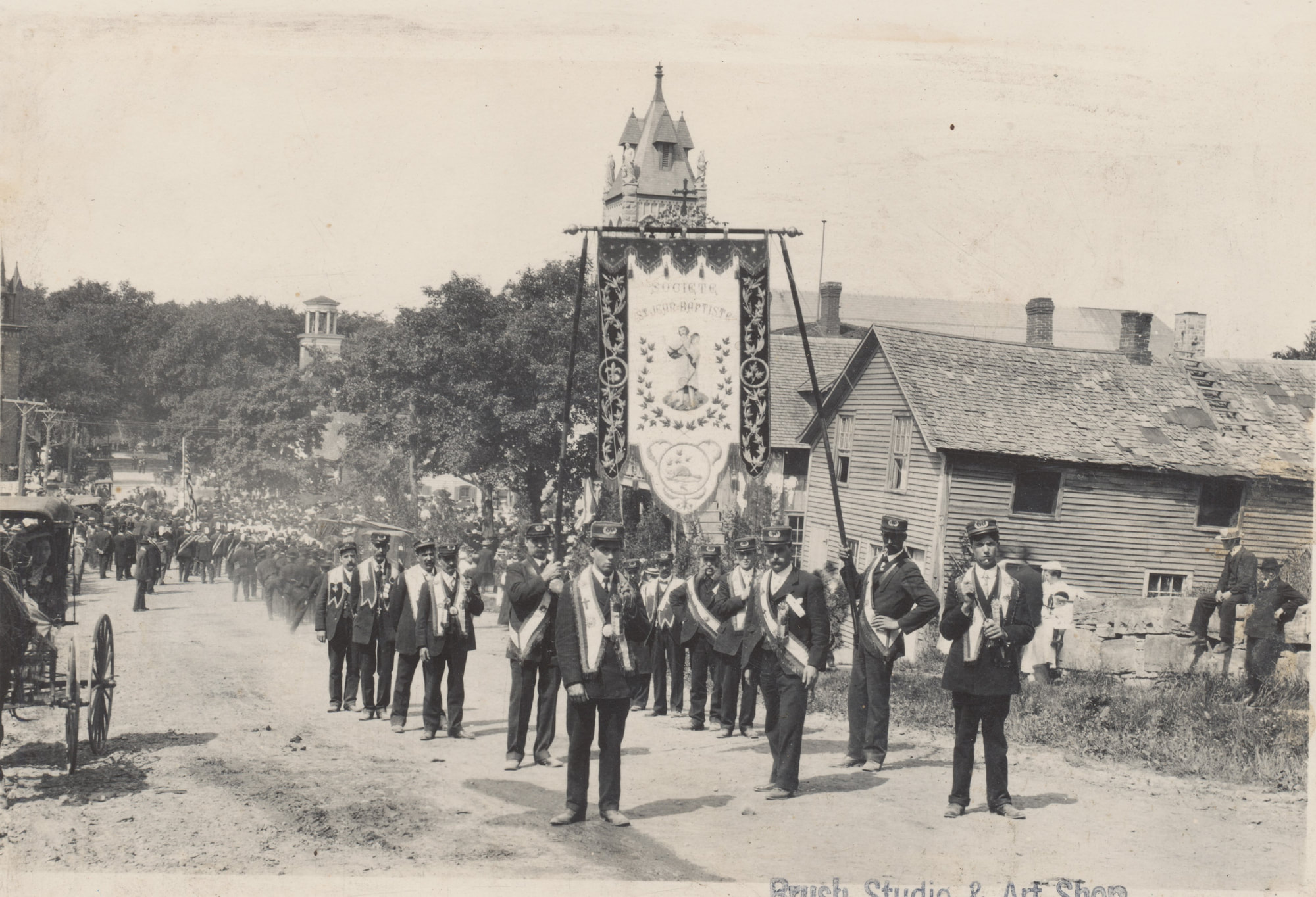 black and white image of people in uniform marching with suspended "Saint-Jean-Baptiste" banner